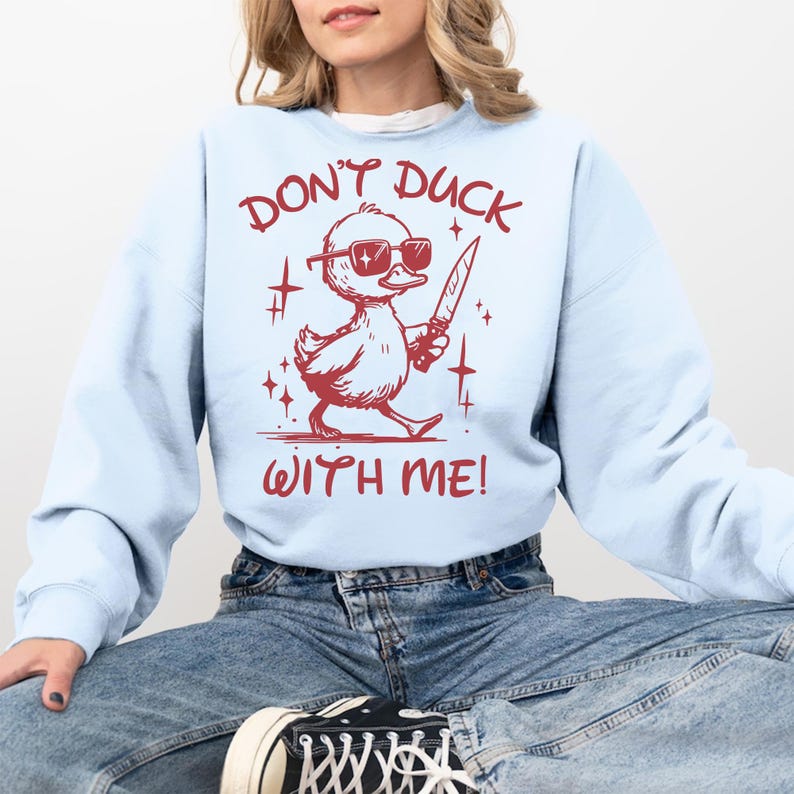 Pode incluir: Uma camisa de frio azul claro com o texto "Don't Duck With Me!" em vermelho. Um pato de desenho animado usando &oacute;culos de sol e segurando uma faca est&aacute; no centro do texto.