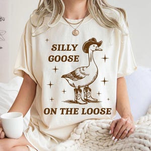 May include: A beige t-shirt with a brown illustration of a goose wearing a cowboy hat and boots. The text "Silly Goose On The Loose" is printed on the shirt.