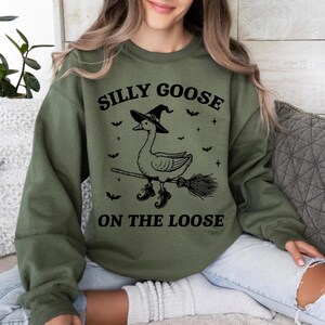 May include: Olive green sweatshirt with a black graphic of a goose wearing a witch hat and boots, riding a broom. The text "SILLY GOOSE ON THE LOOSE" is above and below the goose illustration. Bats and crosses are also included in the design.