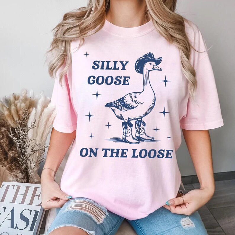 May include: A light pink t-shirt featuring a navy blue illustration of a goose wearing a cowboy hat and boots. The text "SILLY GOOSE ON THE LOOSE" is above and below the goose illustration.