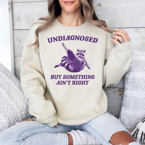 May include: A cream-coloured sweatshirt with a purple graphic of a raccoon doing yoga. The text "UNDIAGNOSED BUT SOMETHING AIN'T RIGHT" is printed on the sweatshirt.