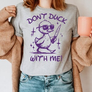 Pode incluir: Uma camiseta cinza com um gr&aacute;fico roxo de um pato usando &oacute;culos de sol e segurando uma faca. O texto "Don't Duck With Me!" est&aacute; impresso na camiseta.