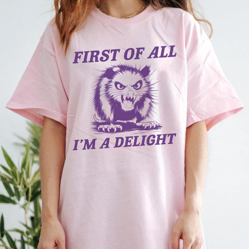 以下が含まれることがあります： 「First of all I'm a delight」というテキストと、オポッサムの紫色のグラフィックが描かれたピンクのTシャツです。