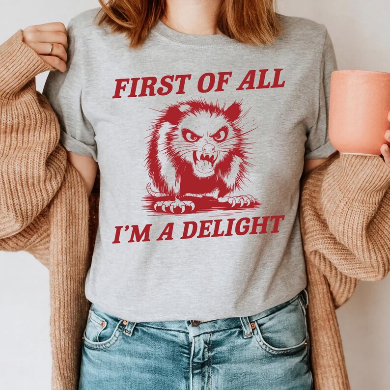 以下が含まれることがあります： 「First of all I'm a delight」というテキストと、赤いオポッサムのグラフィックが描かれたグレーのTシャツ。