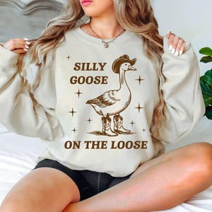 May include: A white sweatshirt with a brown goose wearing cowboy boots and a cowboy hat. The text on the sweatshirt says "Silly Goose On The Loose".