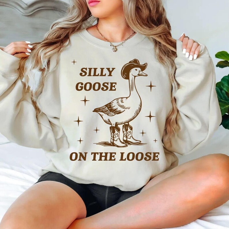 May include: A white sweatshirt with a brown goose wearing cowboy boots and a cowboy hat. The text on the sweatshirt says "Silly Goose On The Loose".