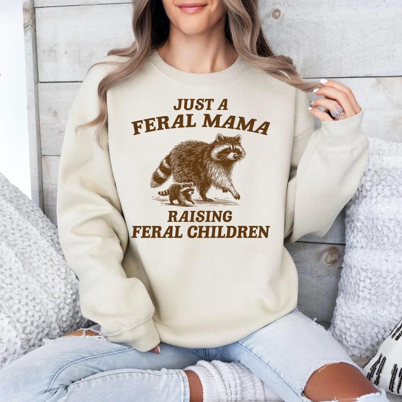 May include: A cream-colored sweatshirt with brown text that reads "Just a Feral Mama Raising Feral Children". A cartoon image of a raccoon and a baby raccoon is printed on the sweatshirt.