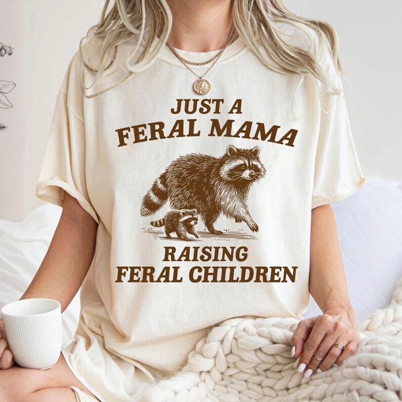 May include: A white t-shirt with the text "Just a Feral Mama Raising Feral Children" printed on it. The shirt features a brown and black raccoon and a smaller raccoon cub.