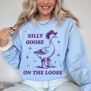 May include: A light blue sweatshirt with a purple graphic of a goose wearing a cowboy hat and boots. The text "Silly Goose On The Loose" is printed on the sweatshirt.