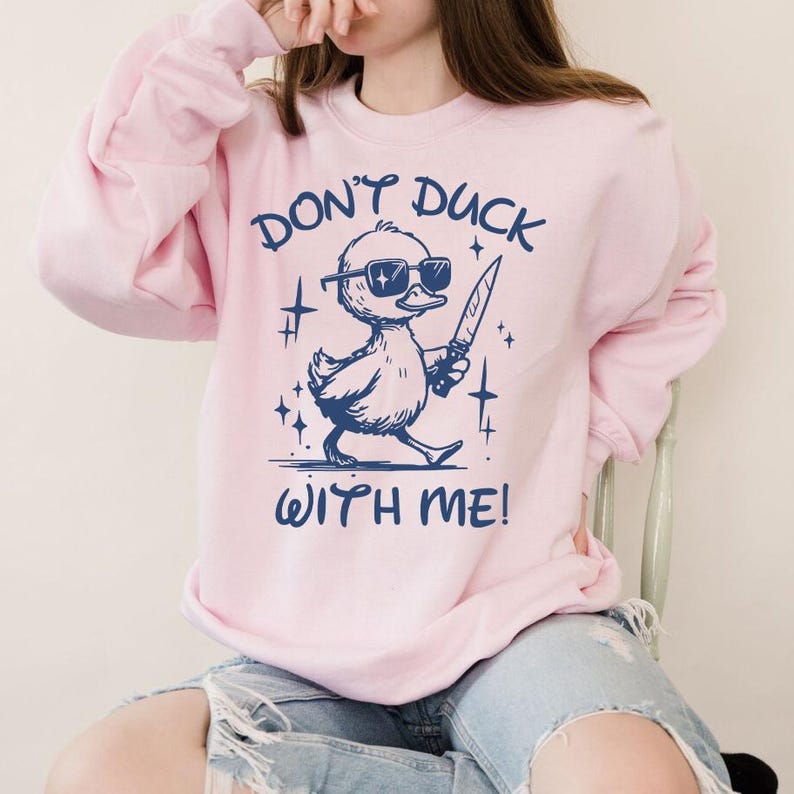 Pode incluir: Uma camisa de moletom rosa com um gr&aacute;fico azul de um pato usando &oacute;culos de sol e segurando uma faca. O texto "Don't duck with me!" est&aacute; impresso abaixo do pato.