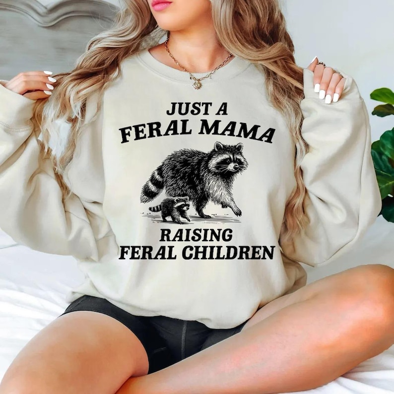 May include: A white sweatshirt with the text "Just a Feral Mama Raising Feral Children" and a black and white illustration of a raccoon and a baby raccoon.
