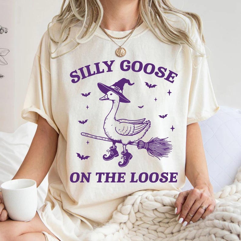 May include: Cream-colored t-shirt with a purple graphic of a goose wearing a witch hat and boots, riding a broom. The text "SILLY GOOSE ON THE LOOSE" is above and below the goose illustration. Bats and stars are also included in the design.