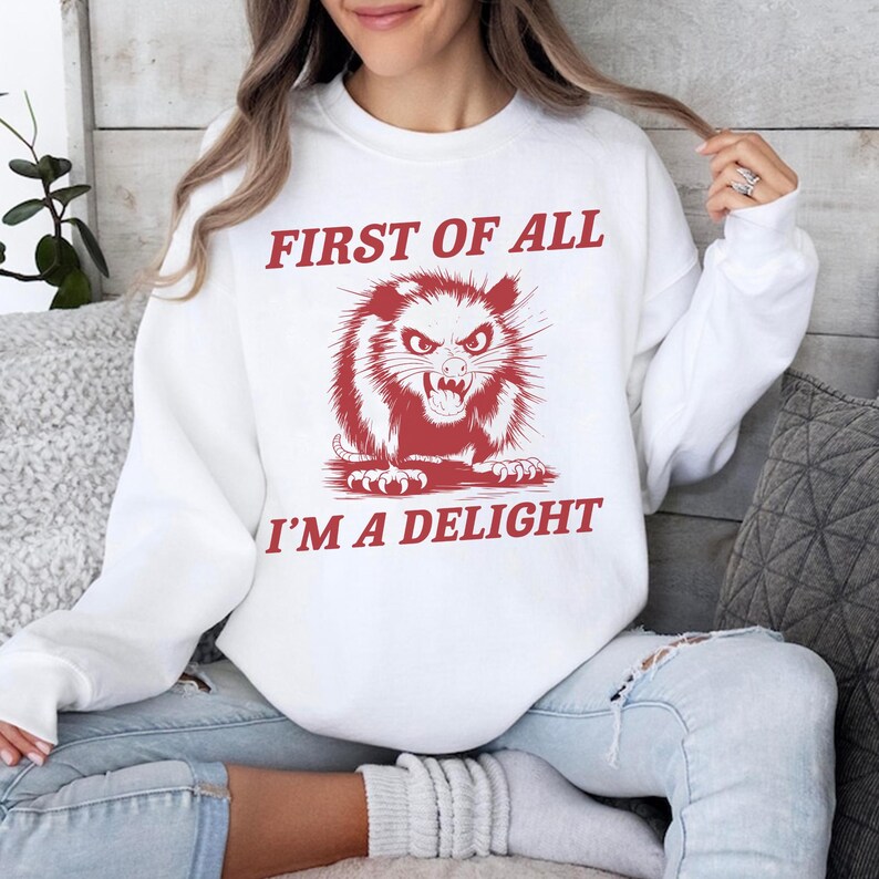 以下が含まれることがあります： 「First of all I'm a delight」という赤い文字が書かれた白いスウェットシャツ。赤い目と赤い口をしたオポッサムの漫画が文字の中央にあります。