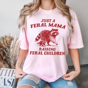 May include: Pink t-shirt with a red graphic of a raccoon and a baby raccoon. The text on the shirt reads "Just a Feral Mama Raising Feral Children."