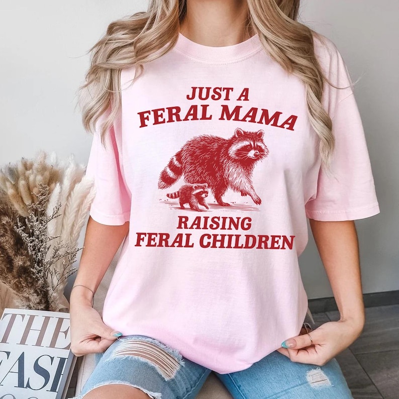 May include: Pink t-shirt with a red graphic of a raccoon and a baby raccoon. The text on the shirt reads "Just a Feral Mama Raising Feral Children."
