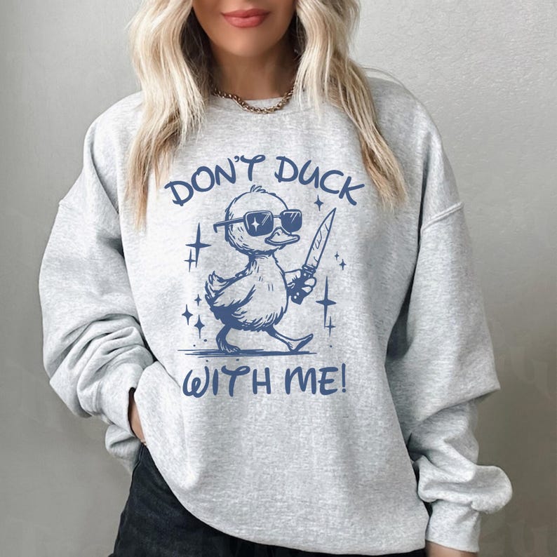 Pode incluir: Uma camisa de moletom cinza com um gr&aacute;fico azul de um pato usando &oacute;culos de sol e segurando uma faca. O texto "Don't duck with me!" est&aacute; impresso abaixo do pato.
