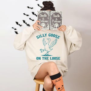 May include: A cream-colored sweatshirt with the text "Silly Goose on the Loose" and a graphic of a goose on roller skates. The person is holding a book with the words "Spells & Potions". Black bat decorations are on the wall.