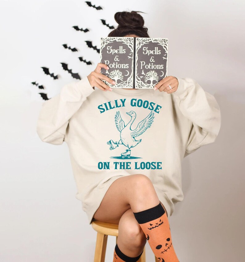 May include: A cream-colored sweatshirt with the text "Silly Goose on the Loose" and a graphic of a goose on roller skates. The person is holding a book with the words "Spells & Potions". Black bat decorations are on the wall.