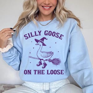May include: Light blue sweatshirt with a purple graphic of a goose wearing a witch hat and boots, riding a broom. The text "SILLY GOOSE ON THE LOOSE" is above and below the goose illustration. The sweatshirt has long sleeves.