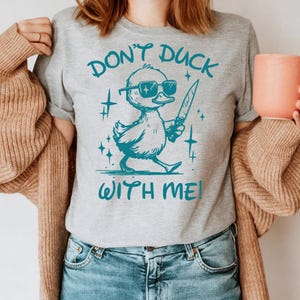 Pode incluir: Camiseta cinza com um gr&aacute;fico turquesa de um pato usando &oacute;culos de sol e segurando uma faca. O texto "Don't Duck With Me!" est&aacute; impresso na camiseta.