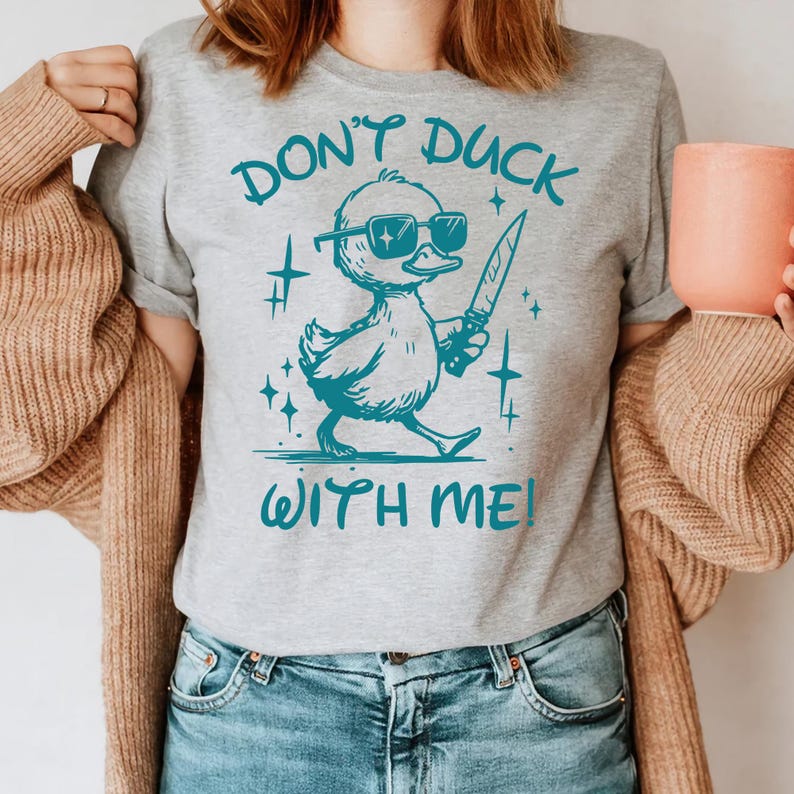 Pode incluir: Camiseta cinza com um gr&aacute;fico turquesa de um pato usando &oacute;culos de sol e segurando uma faca. O texto "Don't Duck With Me!" est&aacute; impresso na camiseta.