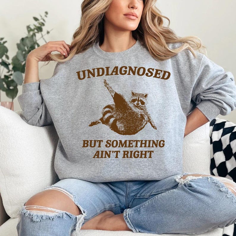 May include: A grey sweatshirt with the text "UNDIAGNOSED BUT SOMETHING AIN'T RIGHT" in brown letters. A cartoon raccoon is pictured in the middle of the text.