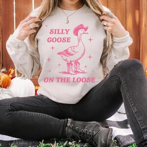 May include: A light gray sweatshirt with a pink graphic of a goose wearing a cowboy hat and boots. The text "Silly Goose On The Loose" is printed on the sweatshirt.