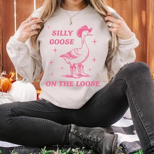 May include: A cream-colored sweatshirt with a pink graphic of a goose wearing a cowboy hat and boots. The text "SILLY GOOSE ON THE LOOSE" is above and below the goose. The sweatshirt is paired with black jeans.