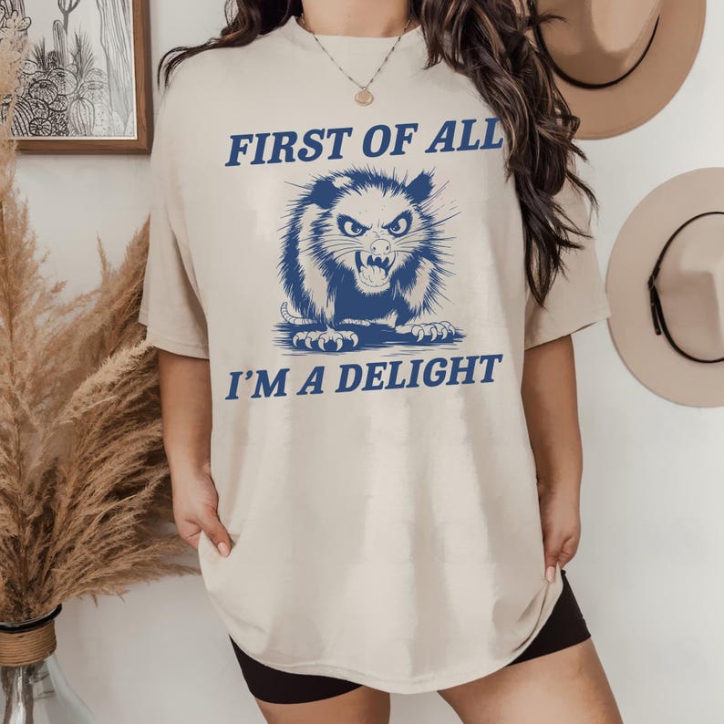 以下が含まれることがあります： ベージュのオーバーサイズTシャツに、オポッサムの青いグラフィックと「First of all I'm a delight」というテキストがプリントされています。