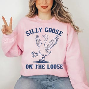May include: A light pink sweatshirt with the words "SILLY GOOSE ON THE LOOSE" in navy blue. The graphic features a goose wearing roller skates. The sweatshirt is a long-sleeved crew neck.