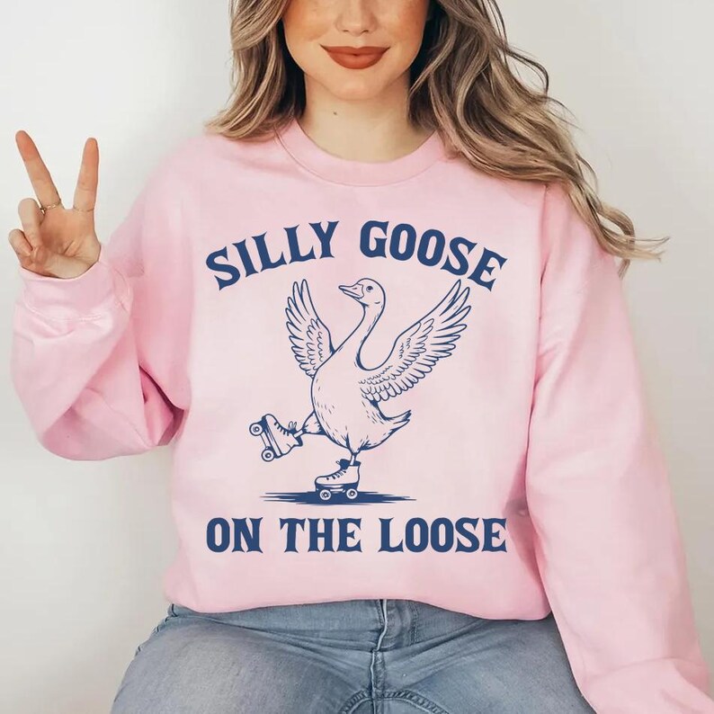 May include: A light pink sweatshirt with the words "SILLY GOOSE ON THE LOOSE" in navy blue. The graphic features a goose wearing roller skates. The sweatshirt is a long-sleeved crew neck.