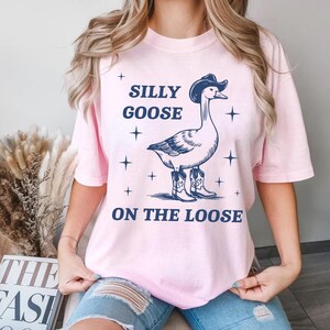 May include: Pink t-shirt with a graphic of a goose wearing a cowboy hat and boots. The text "Silly Goose On The Loose" is printed on the shirt.