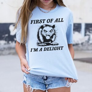 以下が含まれることがあります： 「First of all I'm a delight」というテキストと、オポッサムの白黒イラストが描かれた薄い青色のTシャツです。