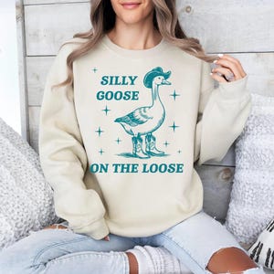 May include: A beige sweatshirt with a teal graphic of a goose wearing a cowboy hat and boots. The text "Silly Goose On The Loose" is printed on the sweatshirt.