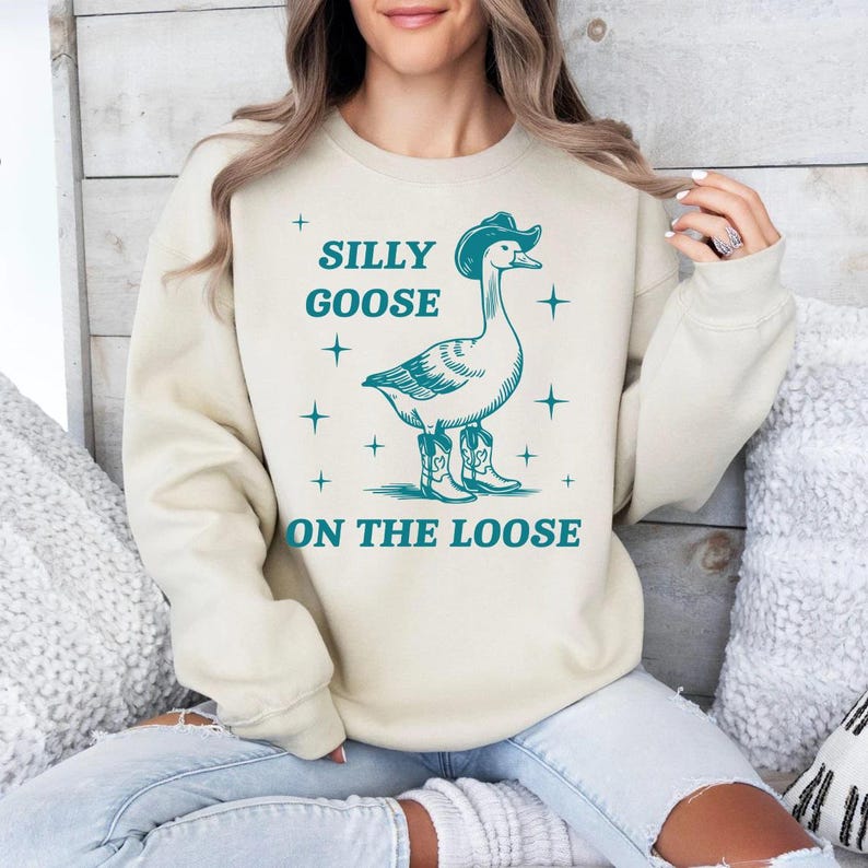 May include: A beige sweatshirt with a teal graphic of a goose wearing a cowboy hat and boots. The text "Silly Goose On The Loose" is printed on the sweatshirt.