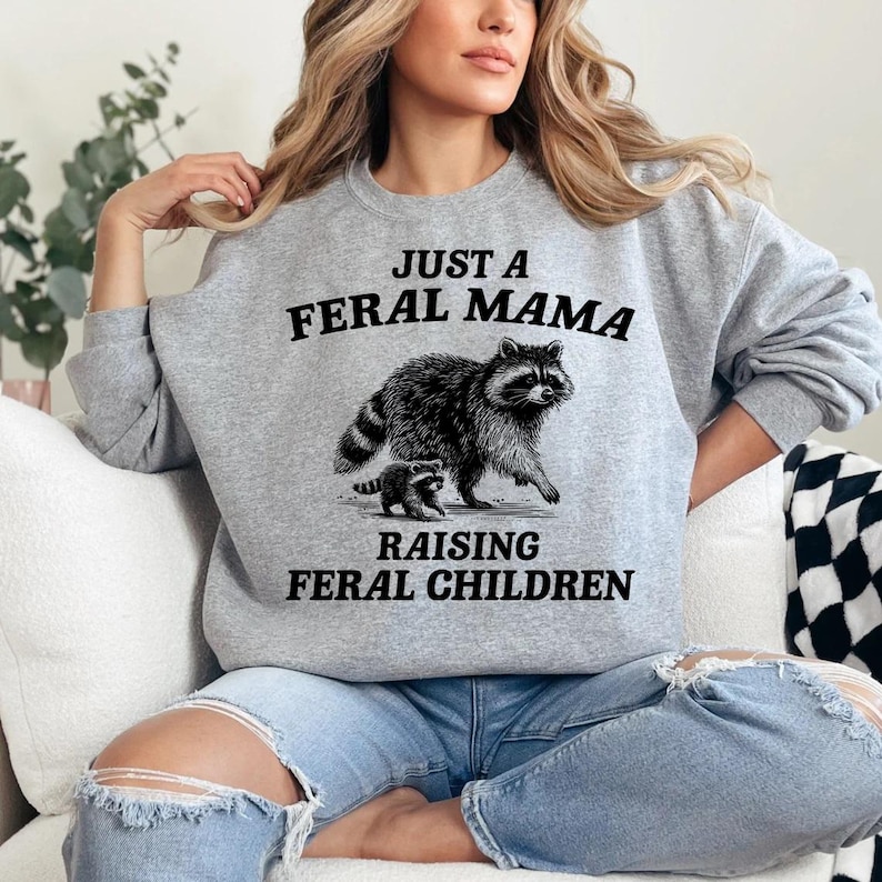 May include: A gray sweatshirt with the text "Just a Feral Mama Raising Feral Children" and a black and white illustration of a raccoon and a baby raccoon.