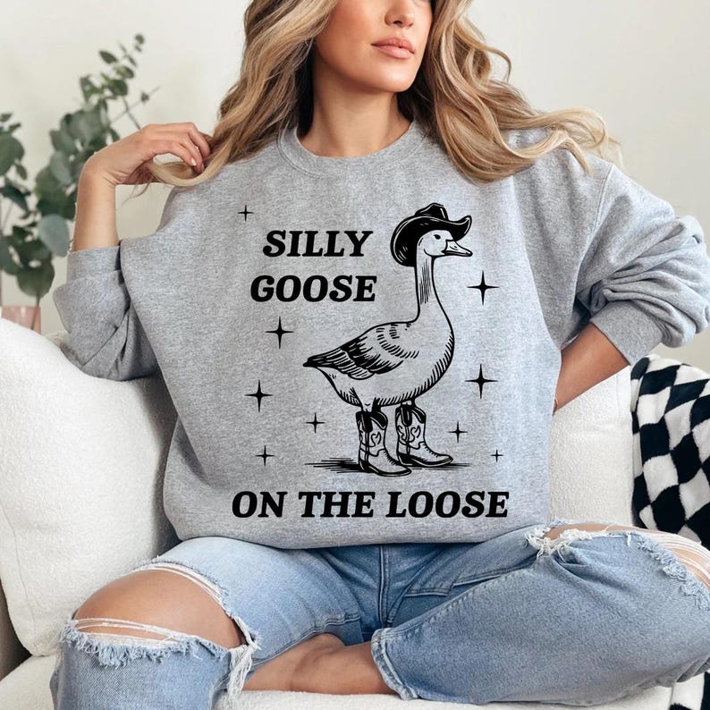 May include: A light gray sweatshirt with a graphic of a goose wearing a cowboy hat and boots. The text "SILLY GOOSE ON THE LOOSE" is printed above and below the goose illustration. The sweatshirt is a casual, comfortable style.
