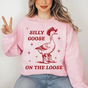 May include: Pink sweatshirt with a red illustration of a goose wearing a cowboy hat and boots. The text "Silly Goose on the Loose" is printed on the sweatshirt.