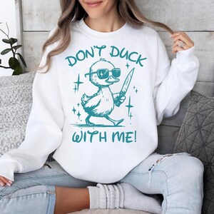 Pode incluir: Camisola branca com um pato turquesa usando &oacute;culos de sol e segurando uma faca. O texto "Don't duck with me!" est&aacute; impresso na camisola.