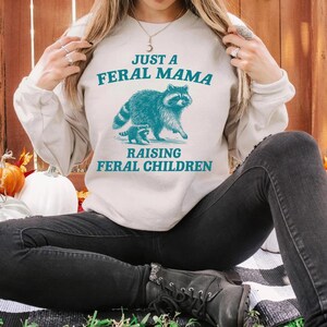 May include: A white sweatshirt with the text "Just a Feral Mama Raising Feral Children" and a blue illustration of a raccoon and a baby raccoon.