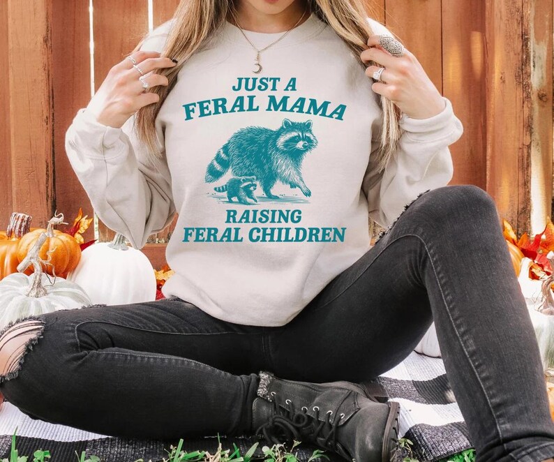 May include: A white sweatshirt with the text "Just a Feral Mama Raising Feral Children" and a blue illustration of a raccoon and a baby raccoon.