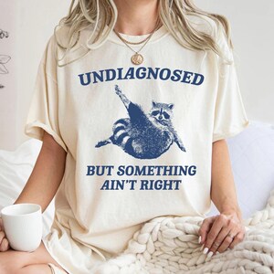 May include: A white t-shirt with a blue raccoon graphic and the text "Undiagnosed but something ain't right".