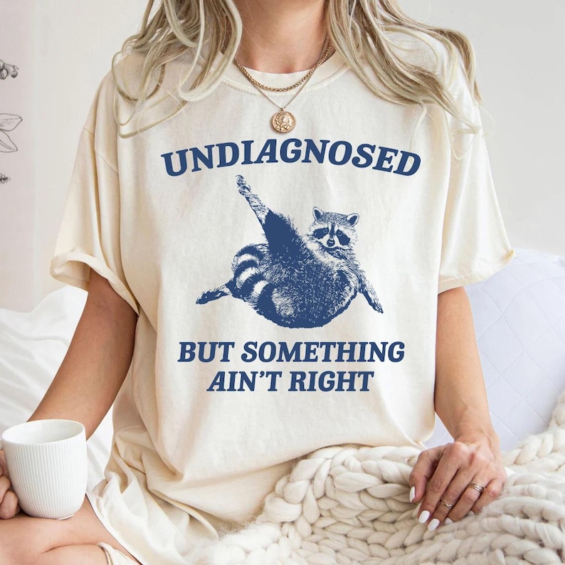 May include: A white t-shirt with a blue raccoon graphic and the text "Undiagnosed but something ain't right".