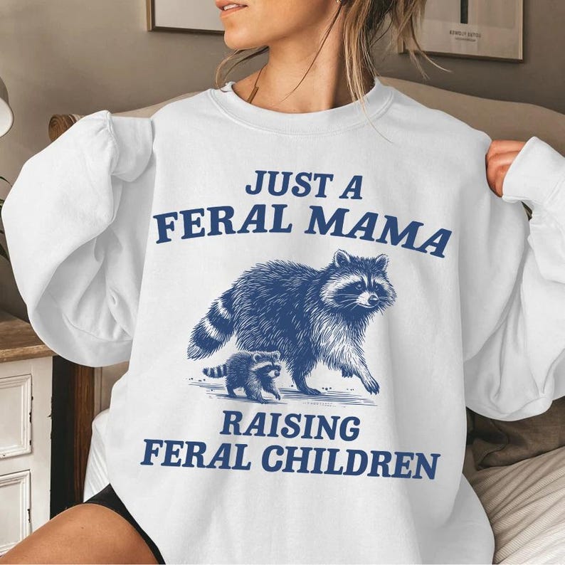 May include: White sweatshirt with a blue graphic of a raccoon and a baby raccoon. The text on the sweatshirt reads "Just a Feral Mama Raising Feral Children."