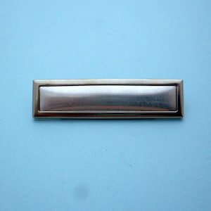 May include: A silver rectangular nameplate with a smooth, reflective surface. The nameplate has a slightly raised border.