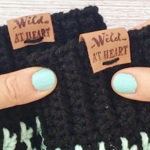 May include: Close-up of two black crocheted items with the text "Wild AT HEART" on brown leather tags. The items have a mint green and white pattern at the bottom. The image includes a person's fingers with light blue nail polish.