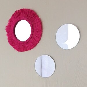 May include: Three round mirrors, one with a pink yarn fringe frame, hanging on a light beige wall.