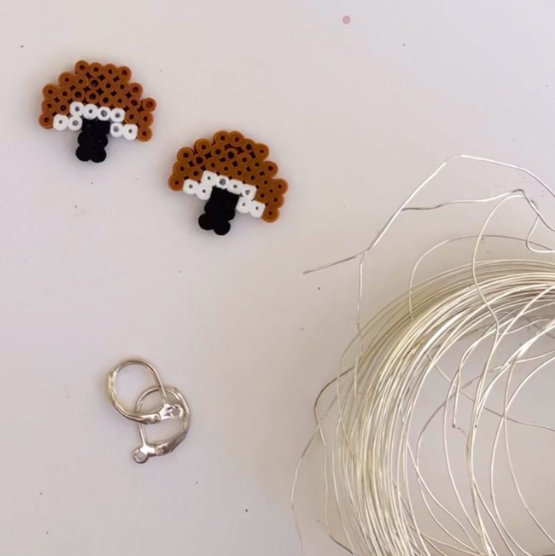 May include: Two mushroom-shaped earrings made from brown, white, and black beads. A silver earring clasp and a coil of silver wire are also present, suggesting a DIY jewelry project. The earrings have a whimsical, handmade aesthetic.
