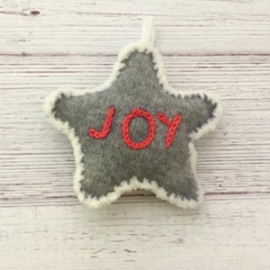 Op de afbeelding: Een grijze viltster-versiering met een witte rand en het woord "JOY" geborduurd in rood.