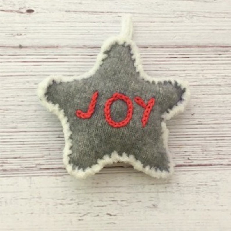Op de afbeelding: Een grijze viltster-versiering met een witte rand en het woord "JOY" geborduurd in rood.